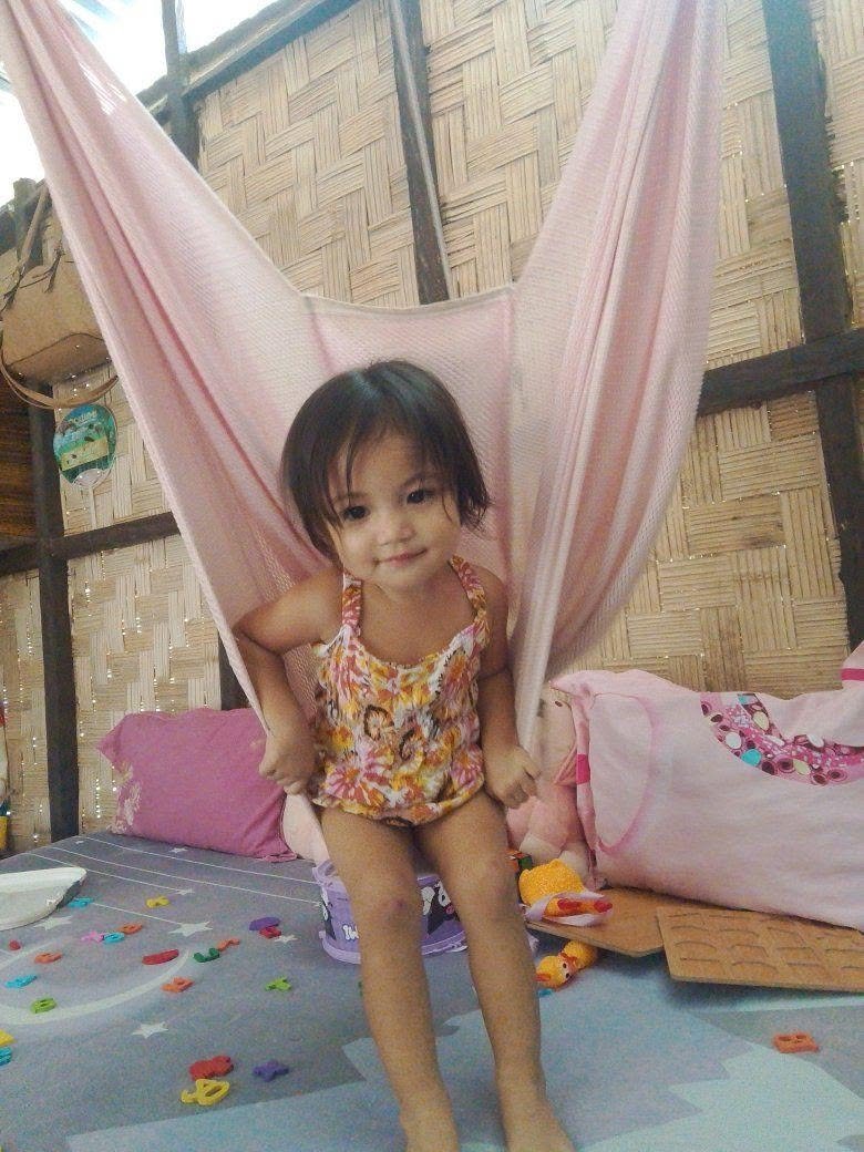 A young girl with short dark hair sitting on a bed in a bamboo-walled room, surrounded by colorful toys and pillows, with a pink hammock or canopy hanging behind her.