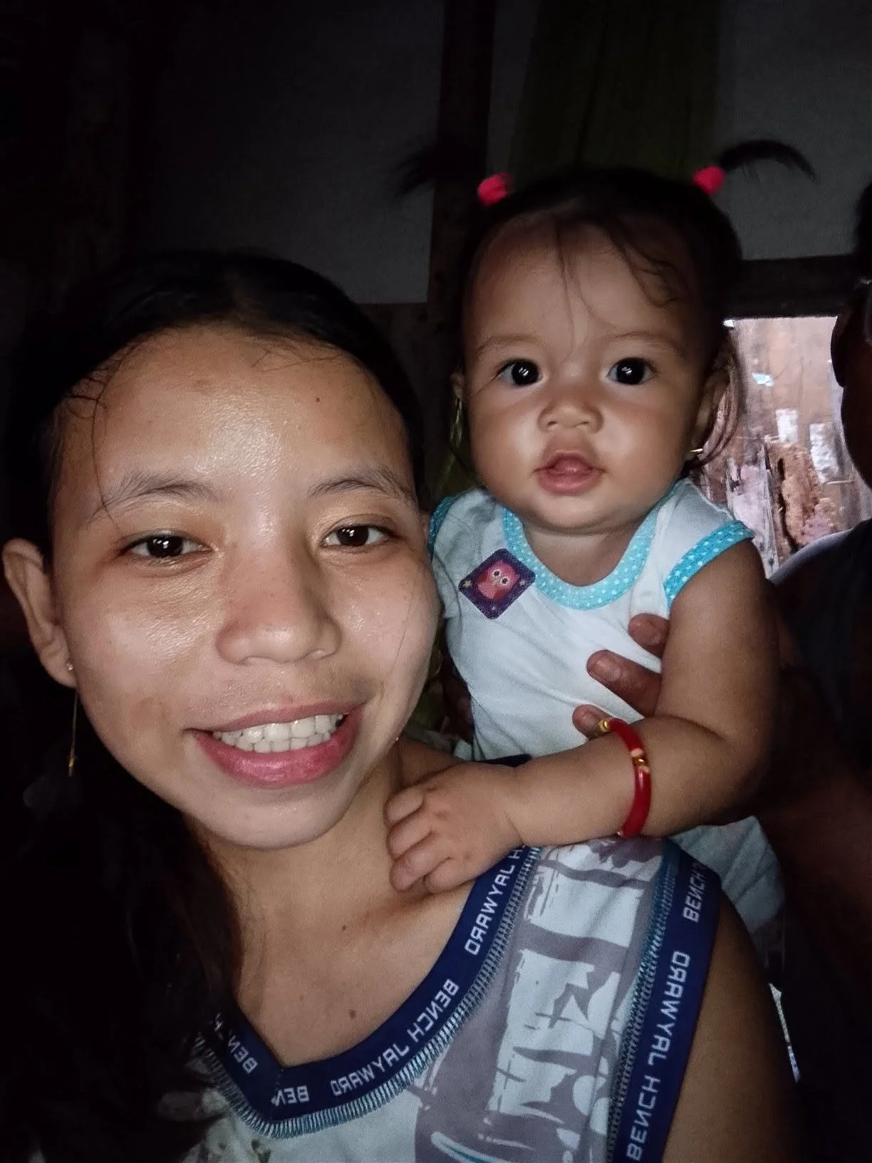 A woman smiling and holding a young girl with pigtails. The girl is looking at the camera and wearing a white top with blue trim, earrings, and a red bracelet. The background appears to be indoors with dark lighting.