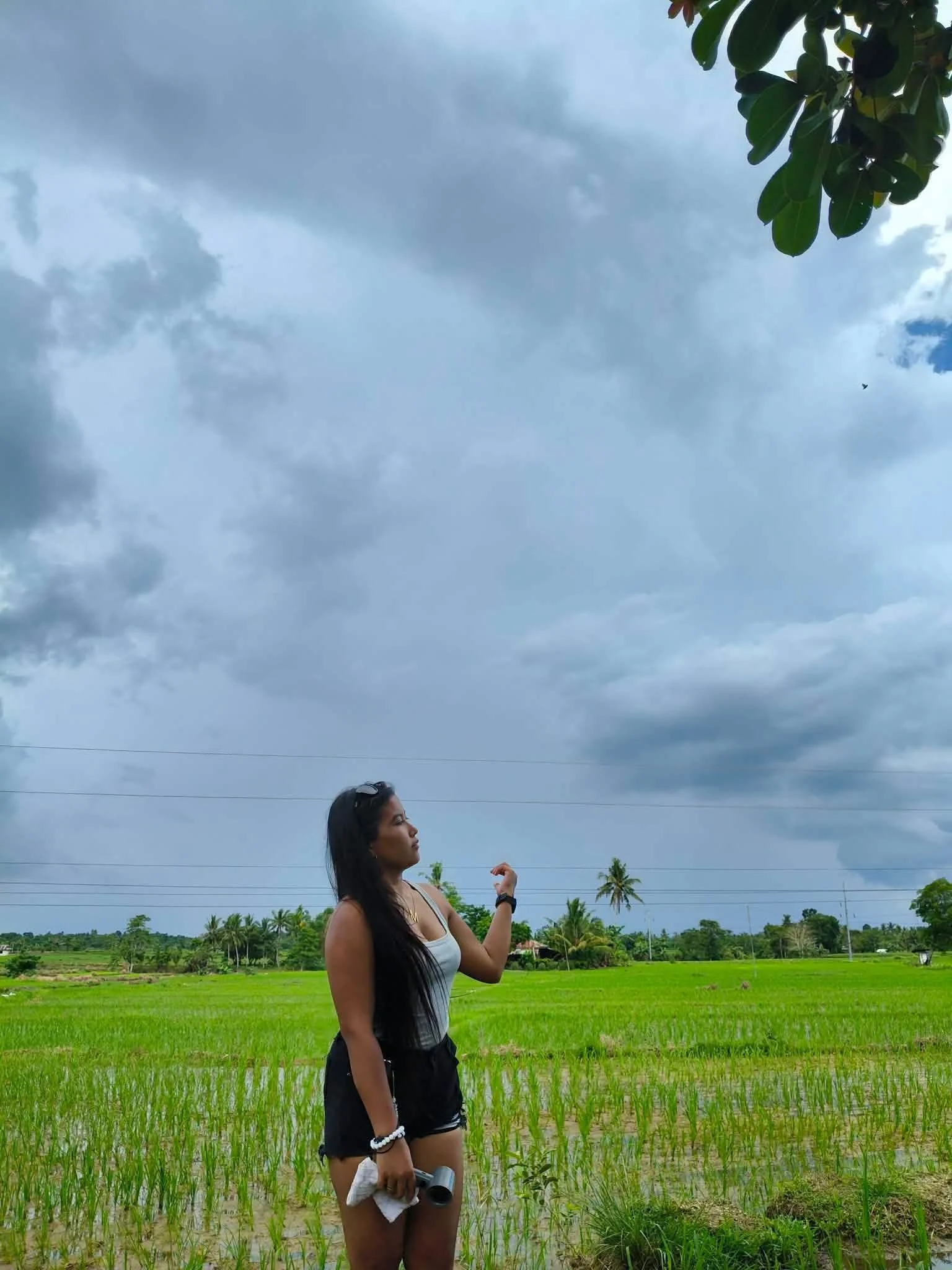 A woman standing in a lush green rice field with a cloudy sky overhead and trees in the background, holding a camera and a towel.