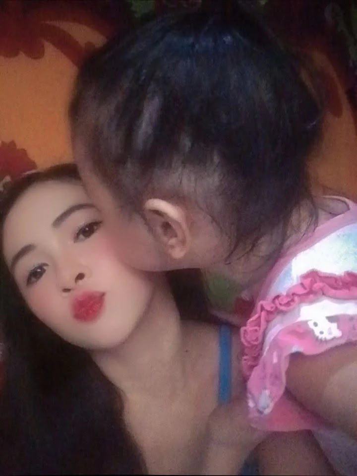 A woman with long hair and makeup making a kiss face while a young girl is giving her a kiss on the cheek, wearing a pink shirt with ruffles.