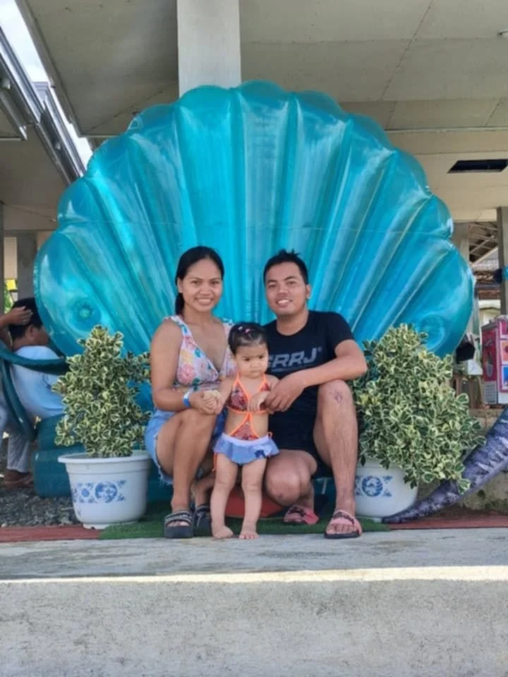 A family of three sitting in front of a large blue seashell sculpture, with green plants in pots on either side, at an outdoor location.