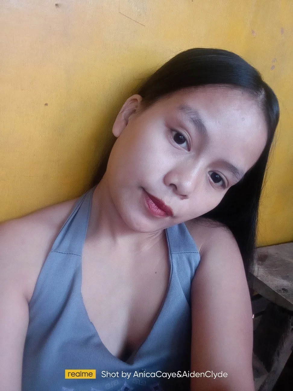 A woman with dark hair and makeup taking a selfie against a yellow wall, wearing a gray sleeveless top.