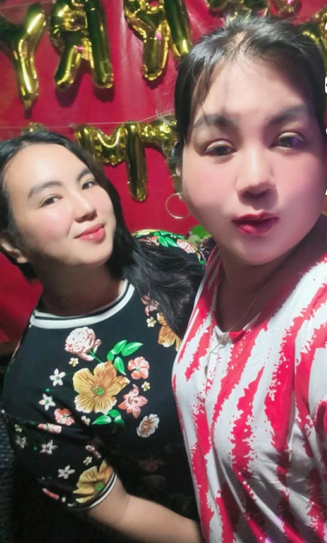 Two women taking a selfie at a celebration with gold balloons in the background. One woman has short black hair, wears hoop earrings and a striped red and white shirt, and is making a slightly surprised expression. The other woman has long dark hair,
