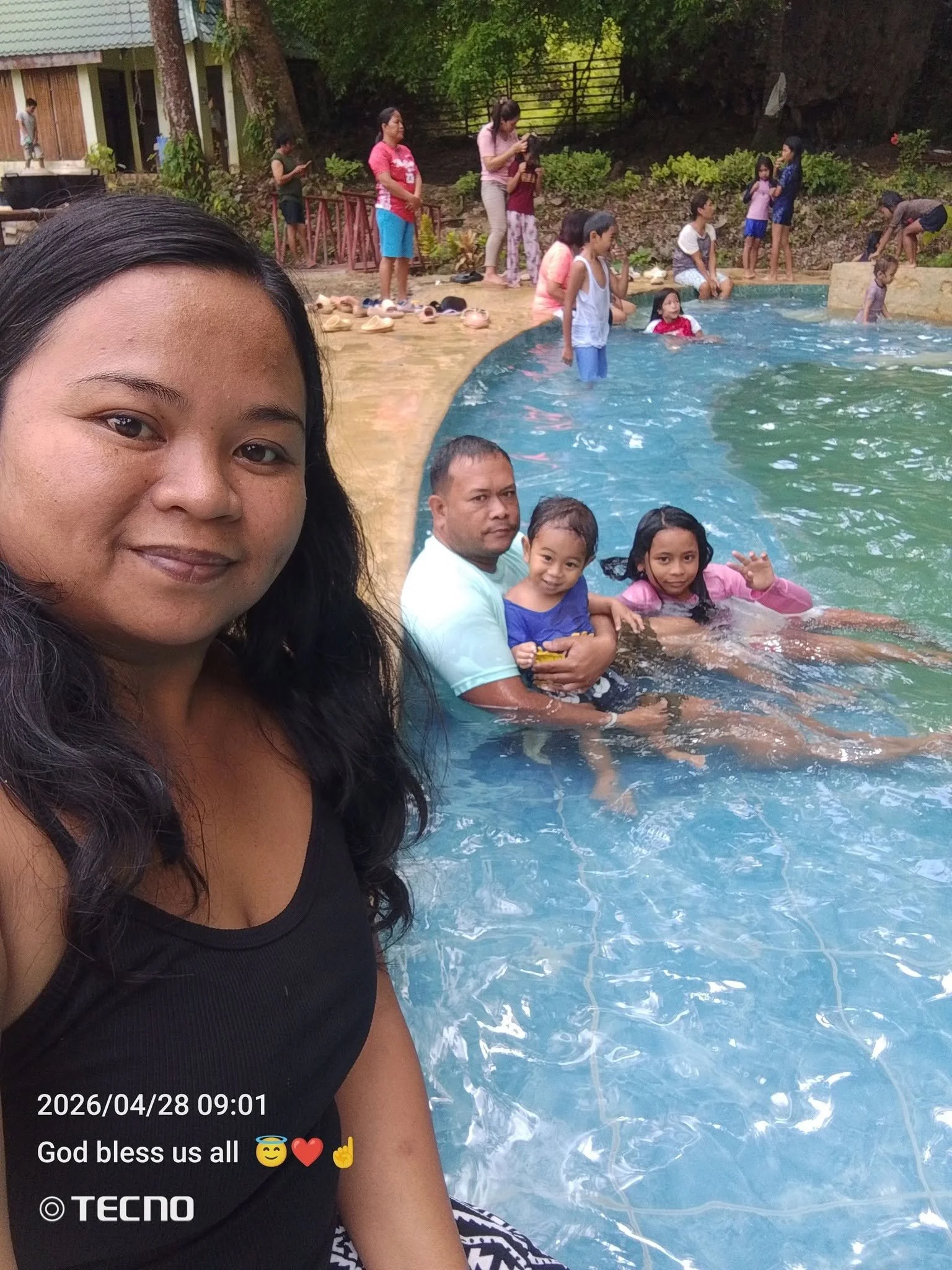 A woman taking a selfie by the poolside with children swimming and playing in the water, while some adults and kids are standing and sitting around the pool area.
