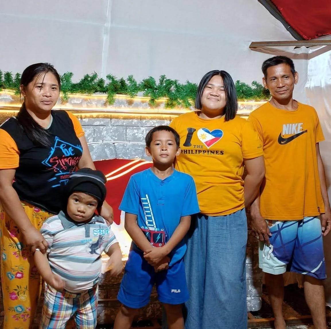 A group of six people, including two children and four adults, standing indoors with green garland and lights in the background. They are wearing casual clothing, some with shirts that have Philippine-themed designs.