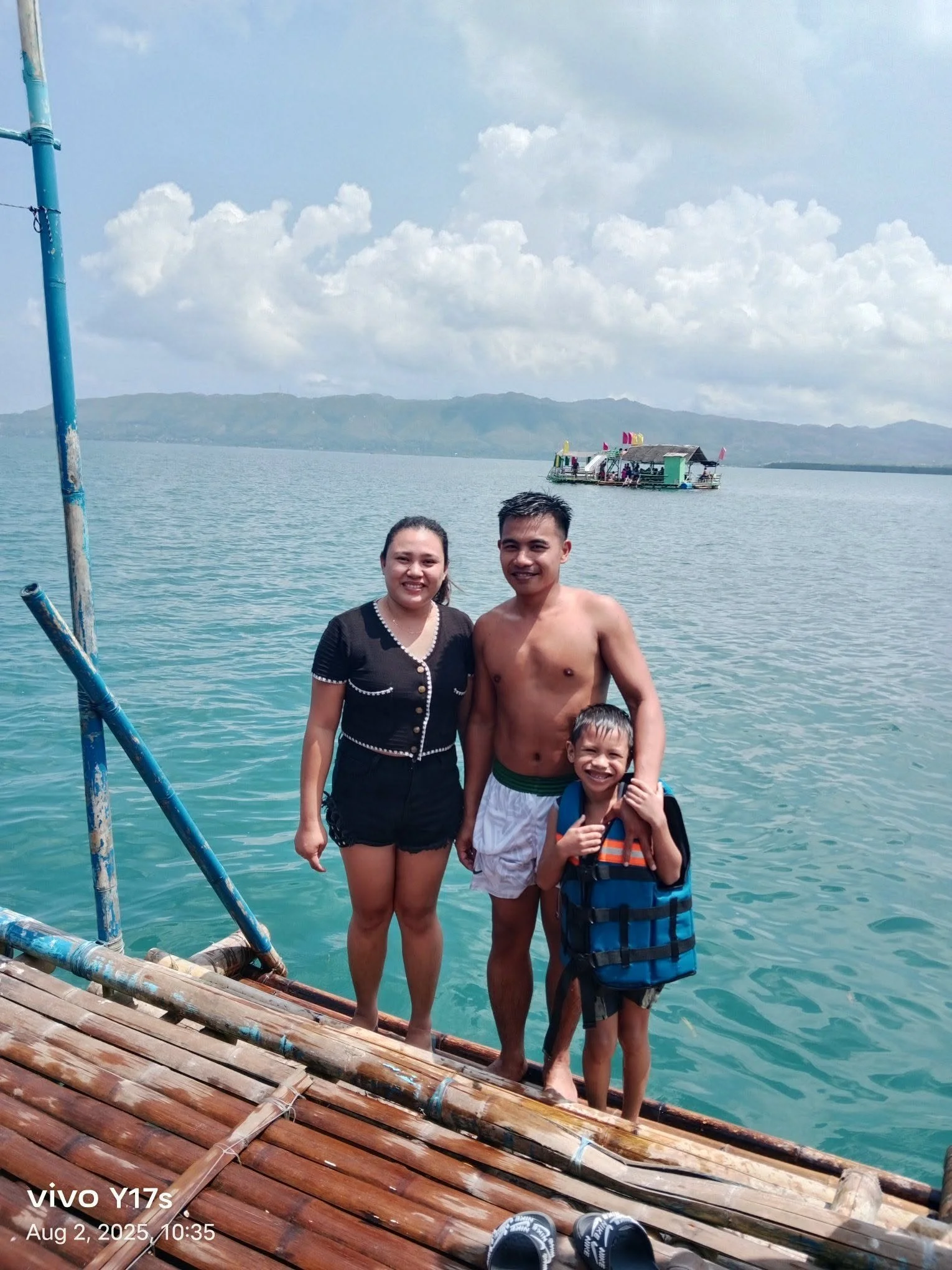 A family standing on a wooden dock by the water, with a floating restaurant or bar in the background and mountains under a partly cloudy sky.