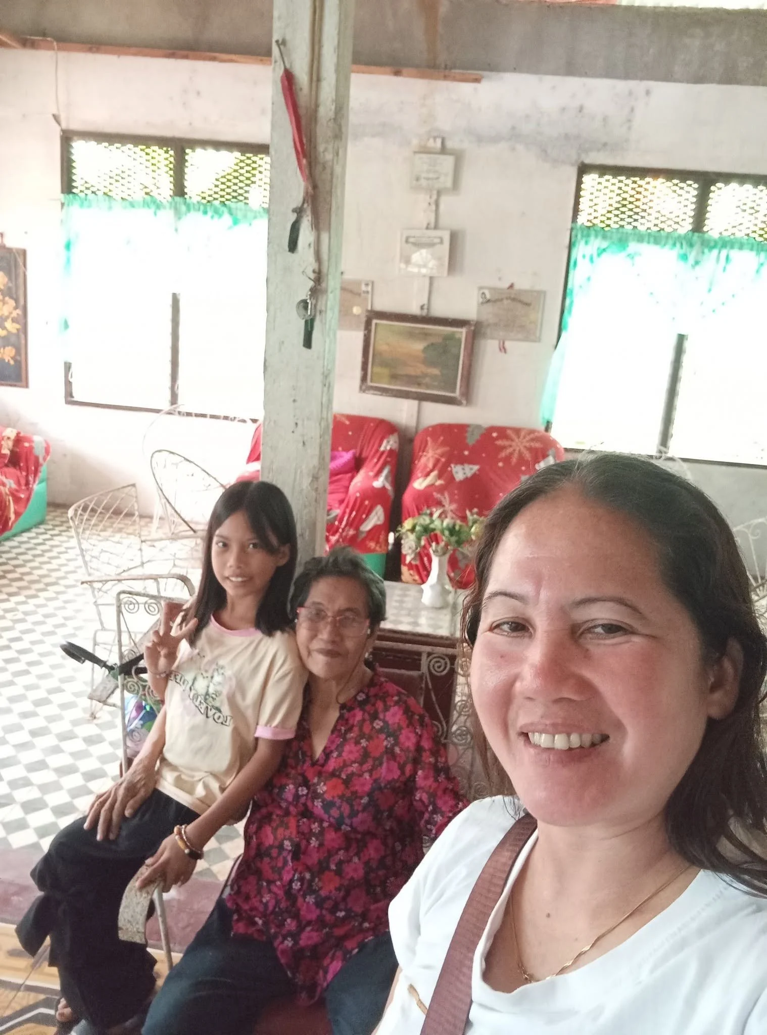 Four women sitting and posing for a photo indoors. Two of them are smiling, and one woman in the foreground is taking the selfie. The background shows a wall with decorations, cushions, and a window with curtains.