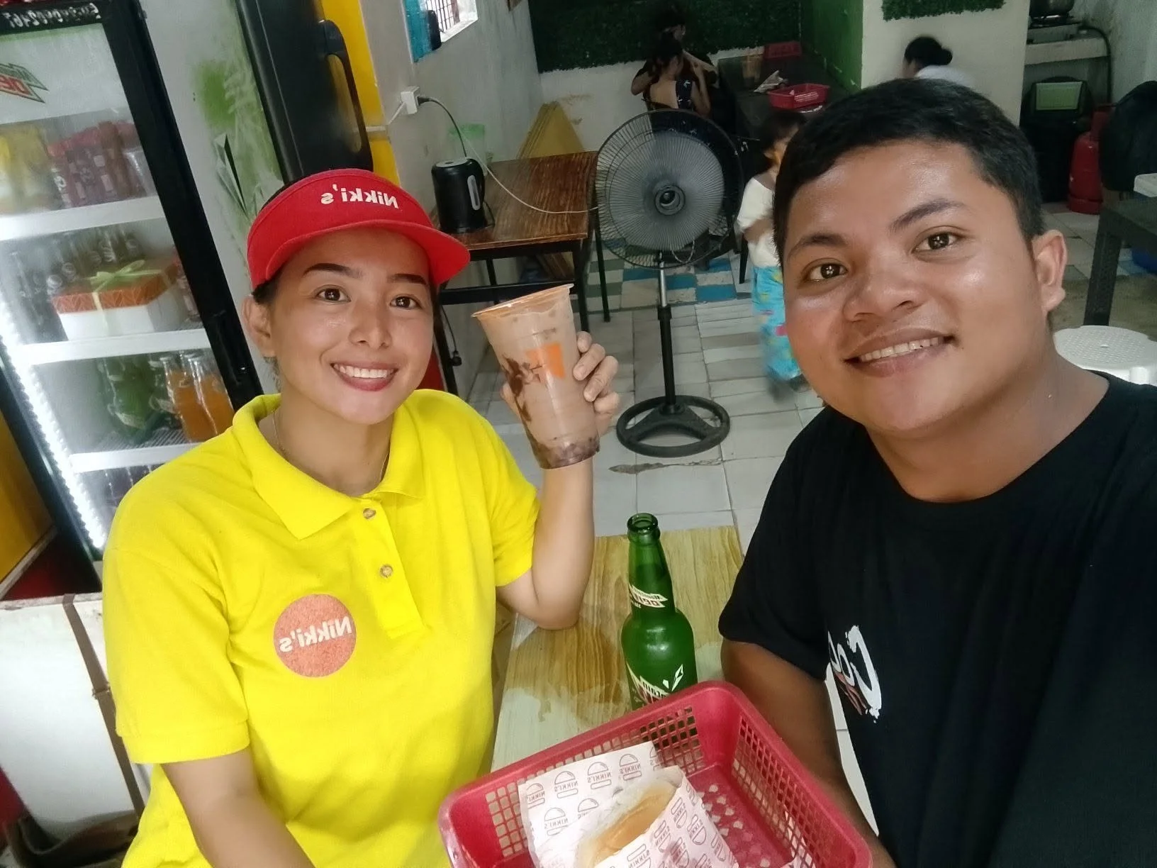 A man and a woman smiling for a selfie at a restaurant. The woman is wearing a yellow shirt and a red cap with 'NIKKI'S' written on it, holding a large iced drink. The man is wearing a black shirt, and a green bottle is on the table. The background s
