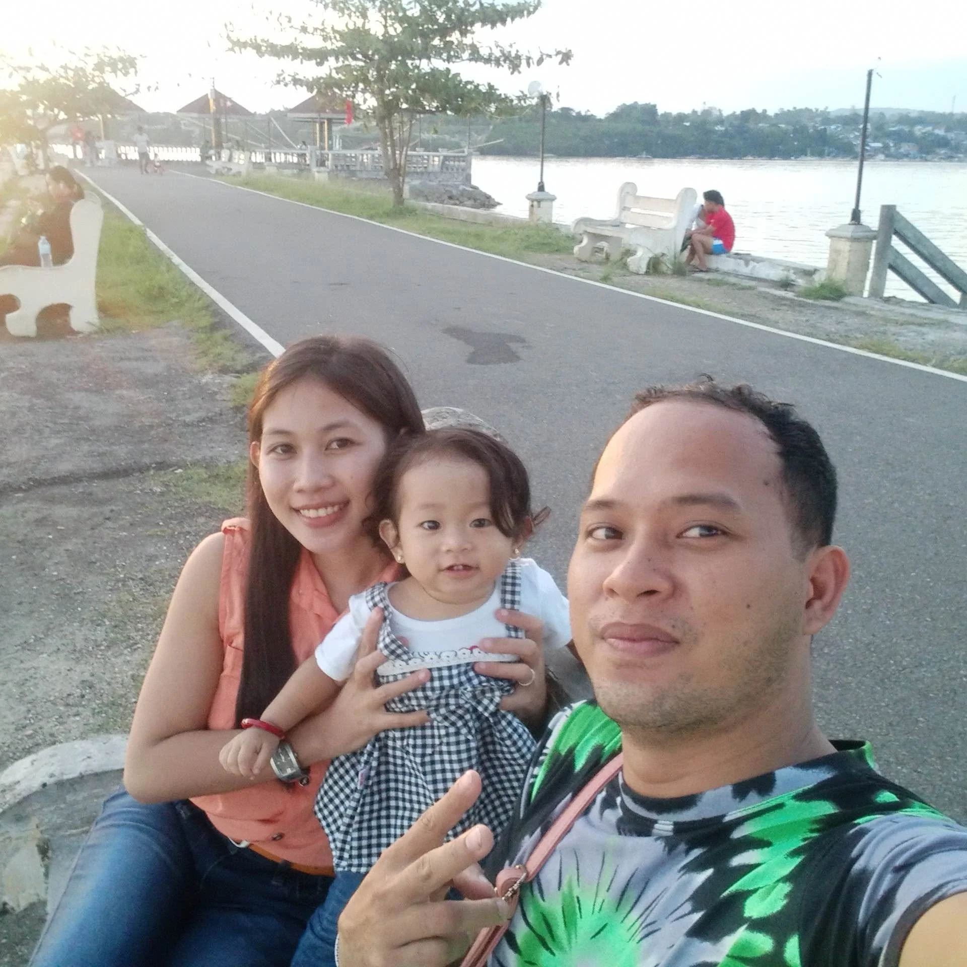 Family taking a selfie near a river with a pedestrian walkway, trees, and benches, during the daytime.