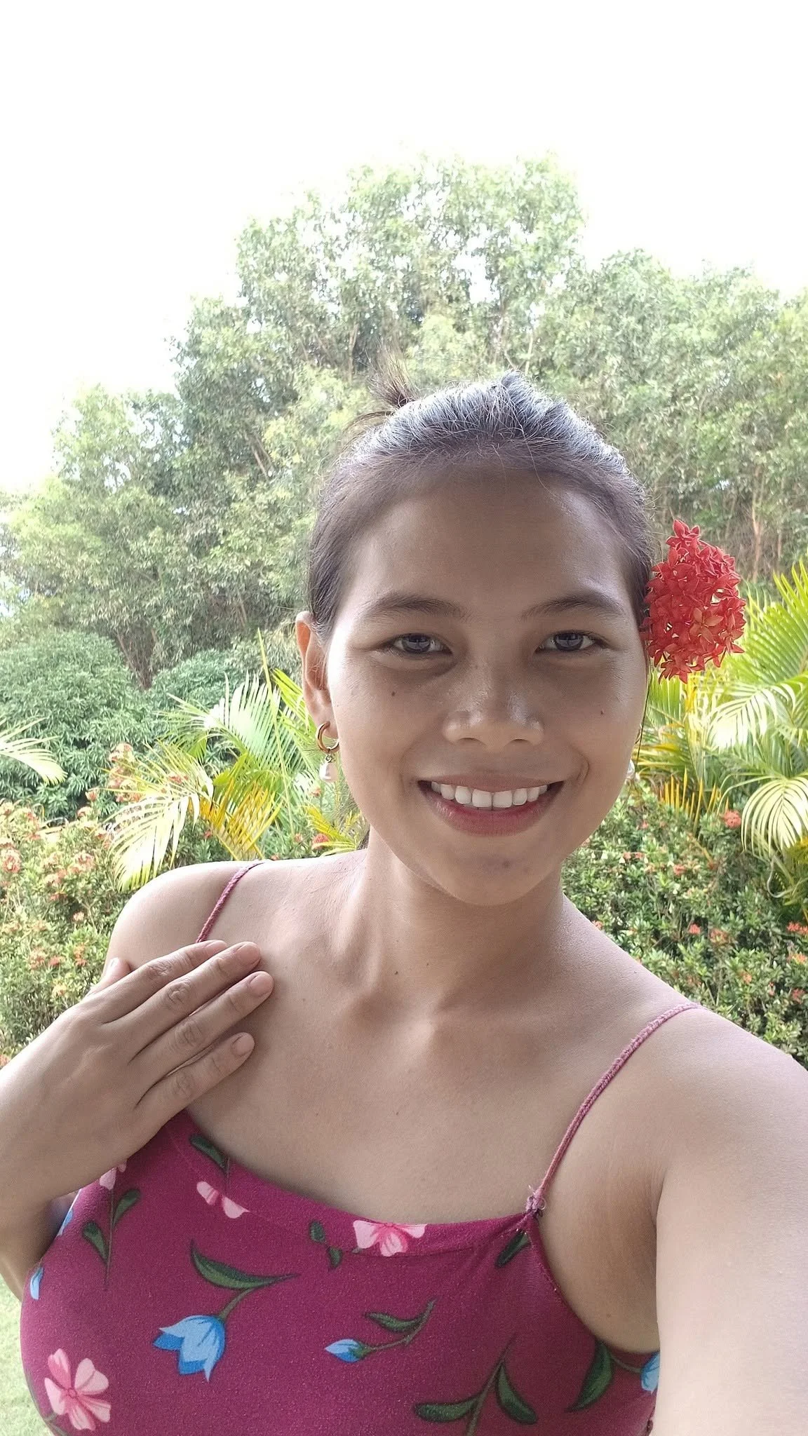 A woman smiling outdoors with lush greenery and trees in the background, wearing a pink floral dress and a red flower in her hair.