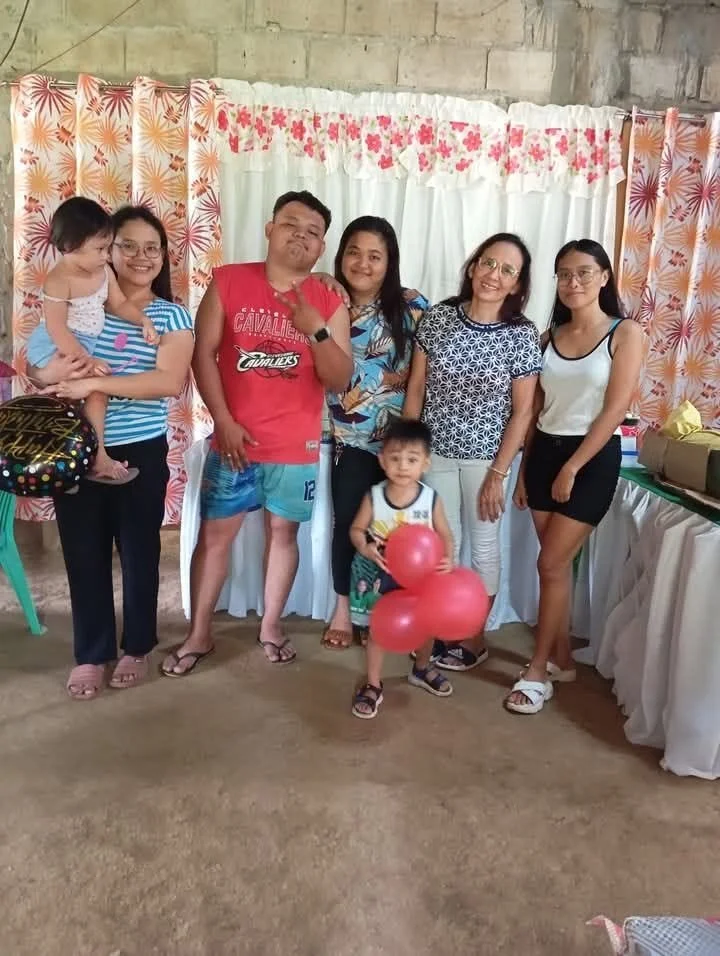 Group of six people, including two children, standing in front of a decorated backdrop with curtains and floral patterns at a celebration or gathering.
