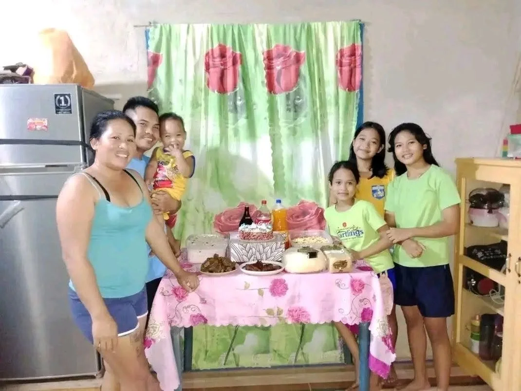 Family gathering in kitchen with a table of food and drinks, women and children smiling.