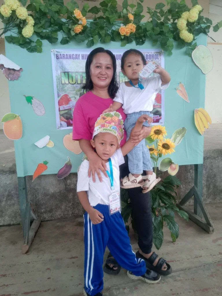 A woman with two children standing in front of a decorated backdrop with fruit and vegetable cutouts. The woman is smiling and wearing a pink shirt, holding one child in her arms, while the other child stands in front of her.