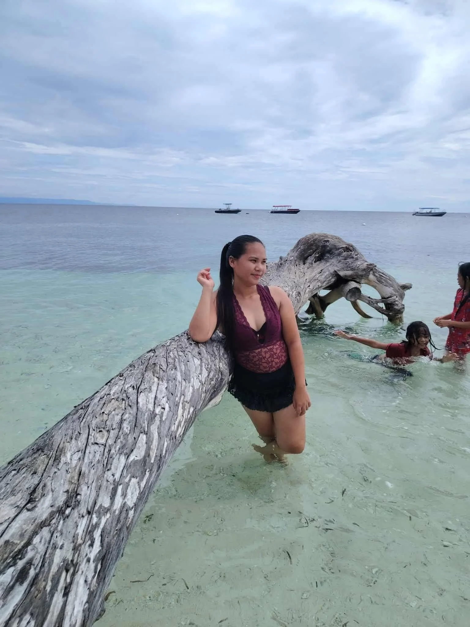 A woman stands in shallow, clear water near a large fallen tree trunk on a beach, with two children playing in the water nearby. There are boats floating in the distance on a cloudy day.