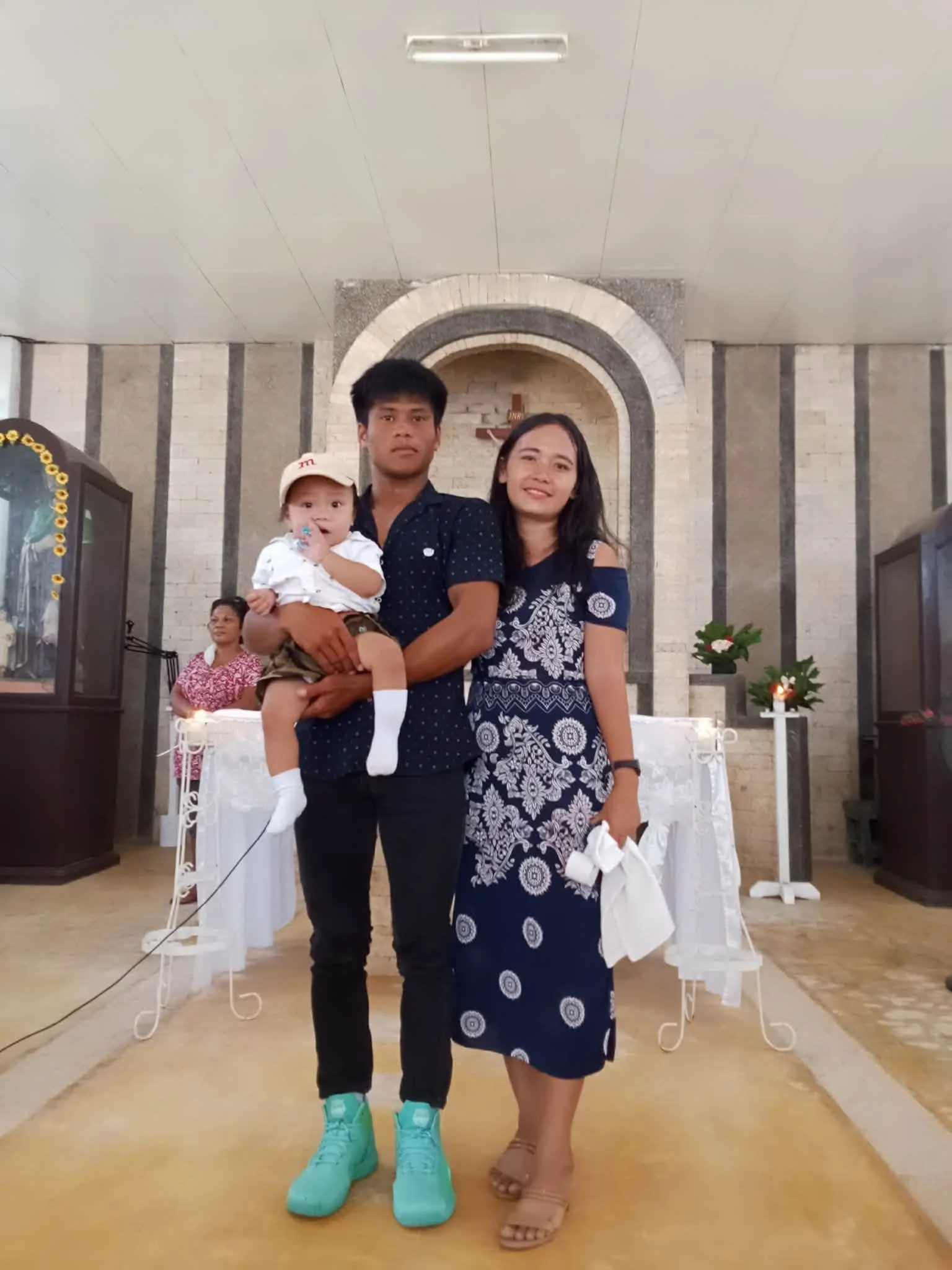 A man holding a young child and a woman standing in a church or chapel. The background features a religious altar with a cross and flowers.