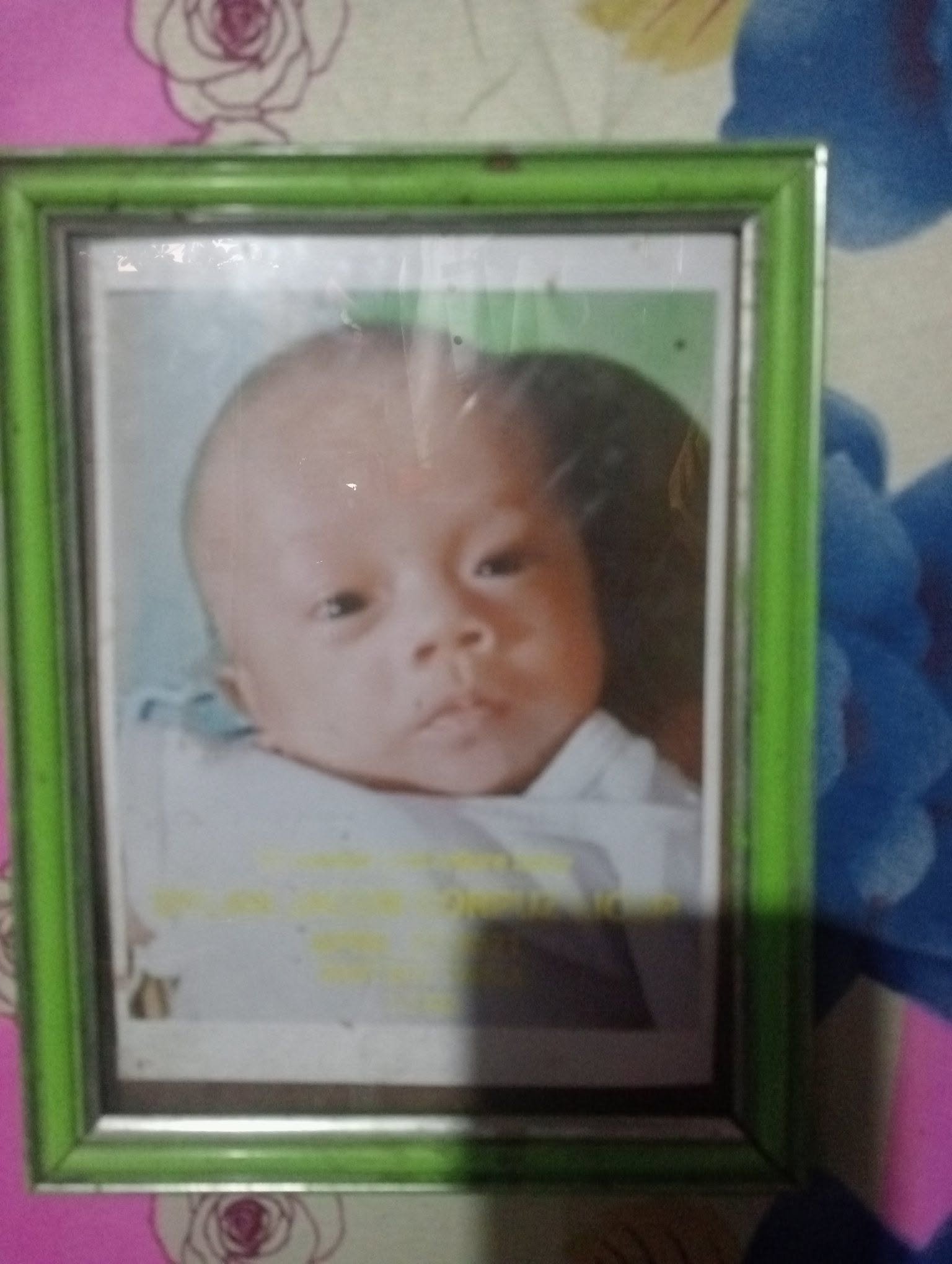 A small rectangular mirror with a green frame reflecting a baby's face with big eyes and a neutral expression, resting on a colorful surface with floral patterns.