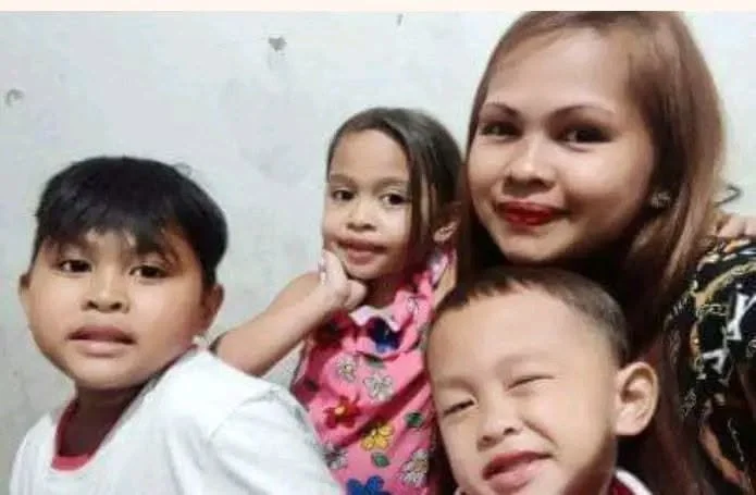 A woman with three children, all smiling and posing for a selfie. The woman has light brown hair and is wearing a patterned top. The children include two boys and a girl, with the girl in the middle wearing a colorful shirt with a butterfly pattern. 