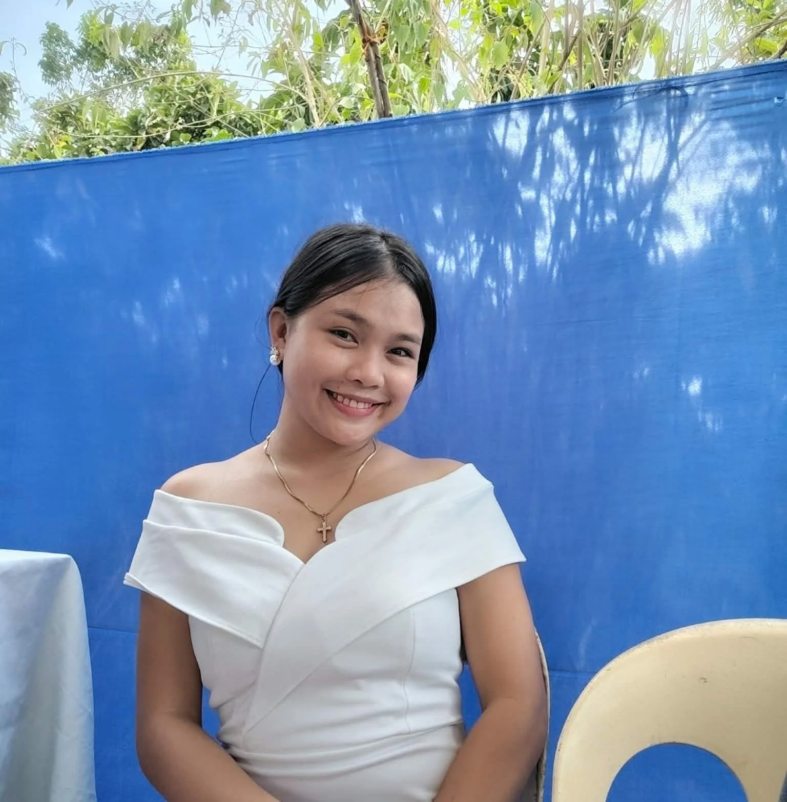 A woman smiling, wearing a white off-shoulder dress, pearl earrings, and a cross necklace, sitting in front of a blue background with trees and sky visible above.
