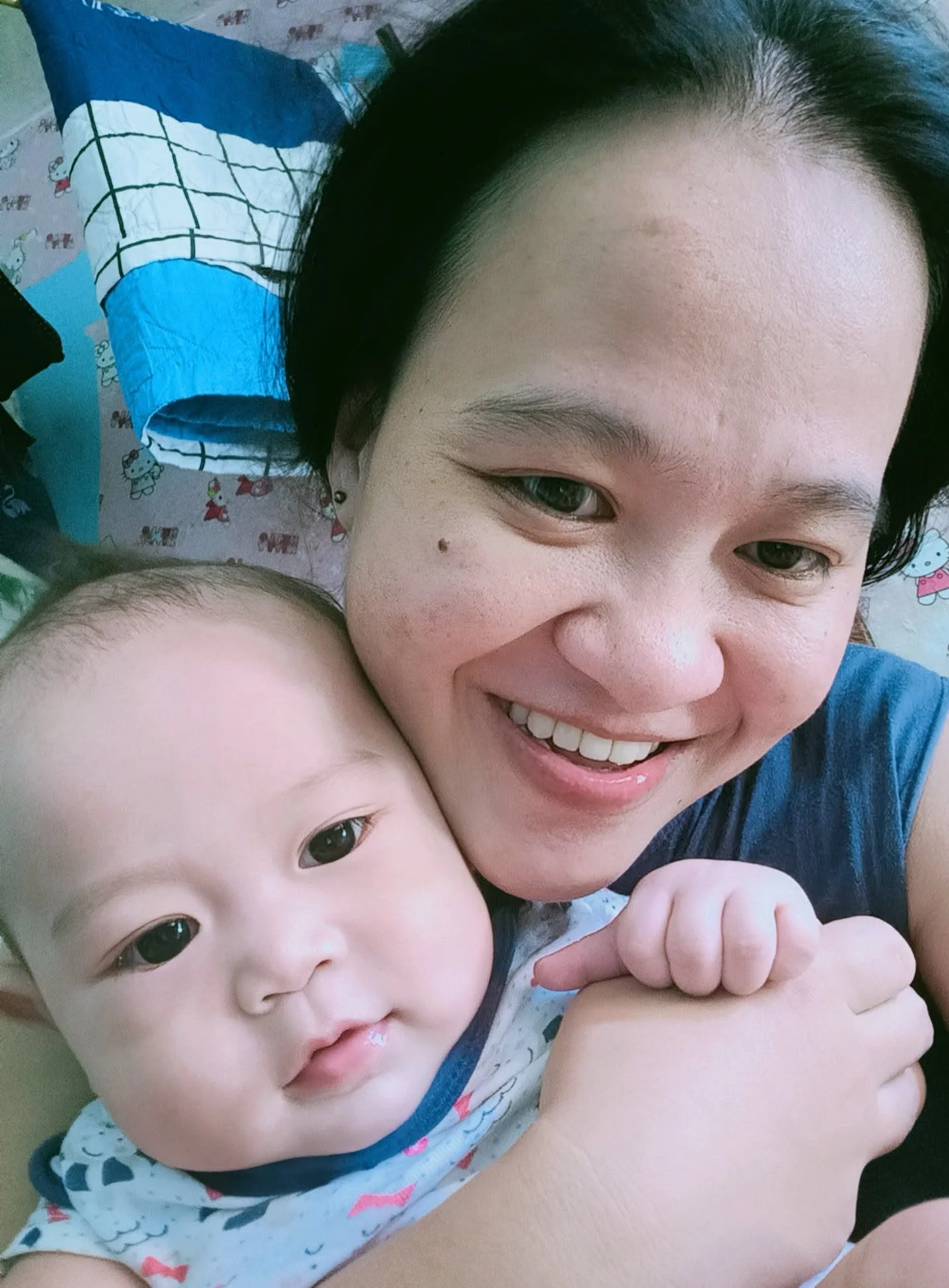 A woman smiling and a baby with big eyes close together, with a background of cartoon-themed bedding.