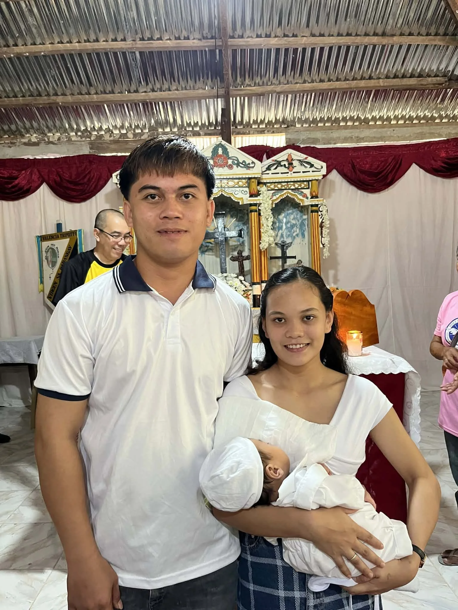 A man and a woman holding a baby, standing inside a church or religious setting with a decorated altar in the background.