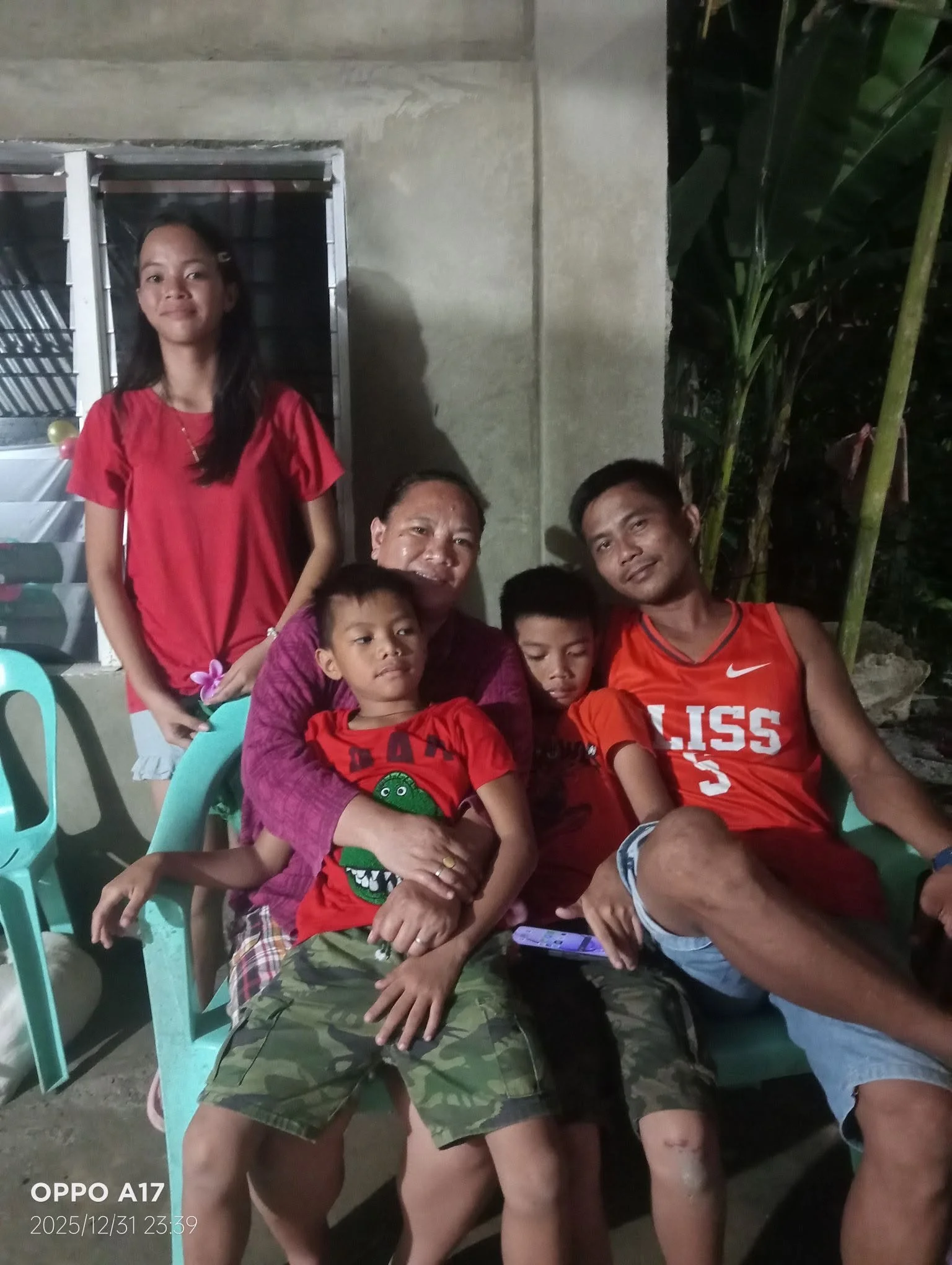 A group of six people, three children and three adults, gathered outdoors at night, some sitting on plastic chairs, with a concrete wall and greenery in the background.