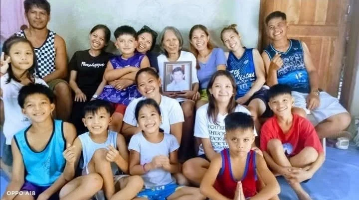 Group of children and an older woman sitting indoors, holding a framed photograph, smiling for the camera.