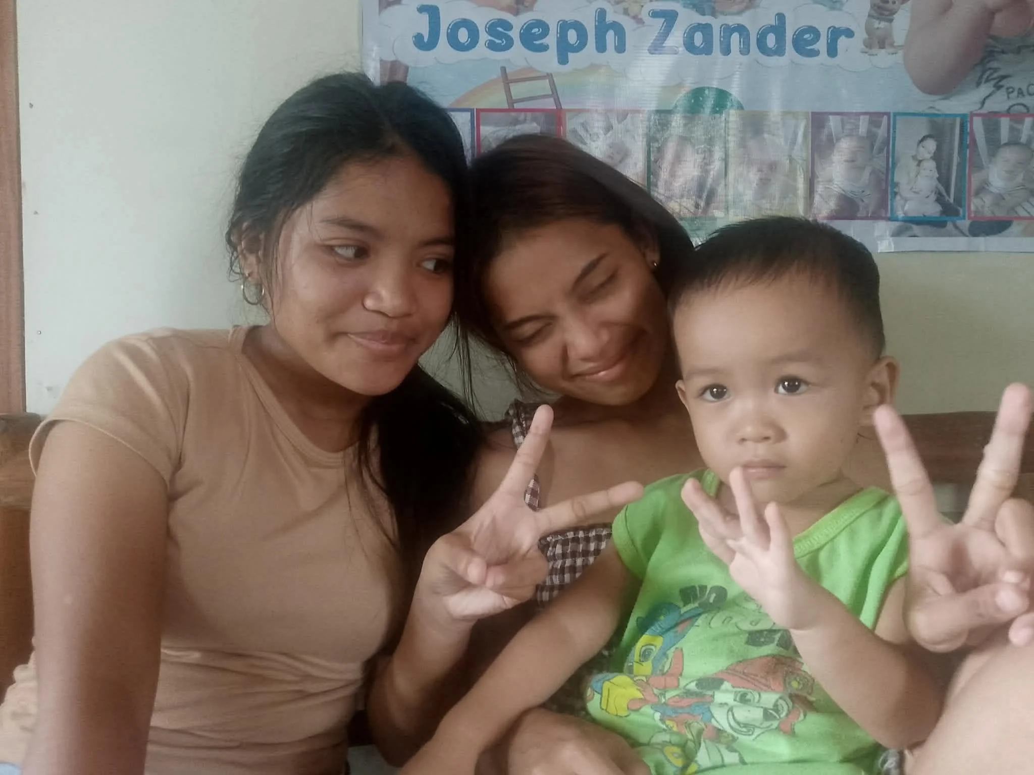 Three people, two women and a young boy, close together, making peace signs with their fingers. The background includes a colorful poster with the name 'Joseph Zander' and cartoon characters.