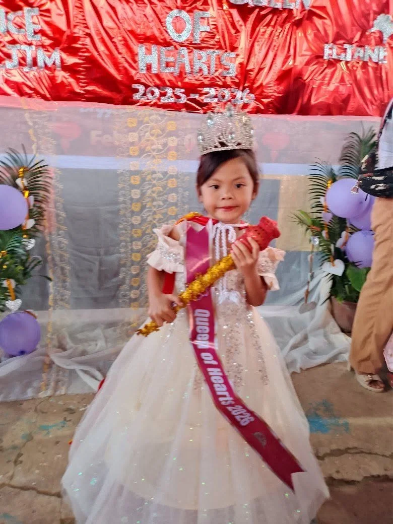 Young girl dressed as a queen or princess, wearing a crown and a white gown, holding a royal scepter, standing in front of a decorated backdrop with balloons and greenery at a celebration.