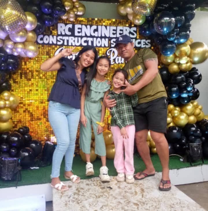 Four people, two adults and two children, standing and smiling in front of a decorated backdrop with gold, black, and purple balloons, and a sign that reads 'RC Engineers Construction Services.' The scene appears to be a celebration or party.