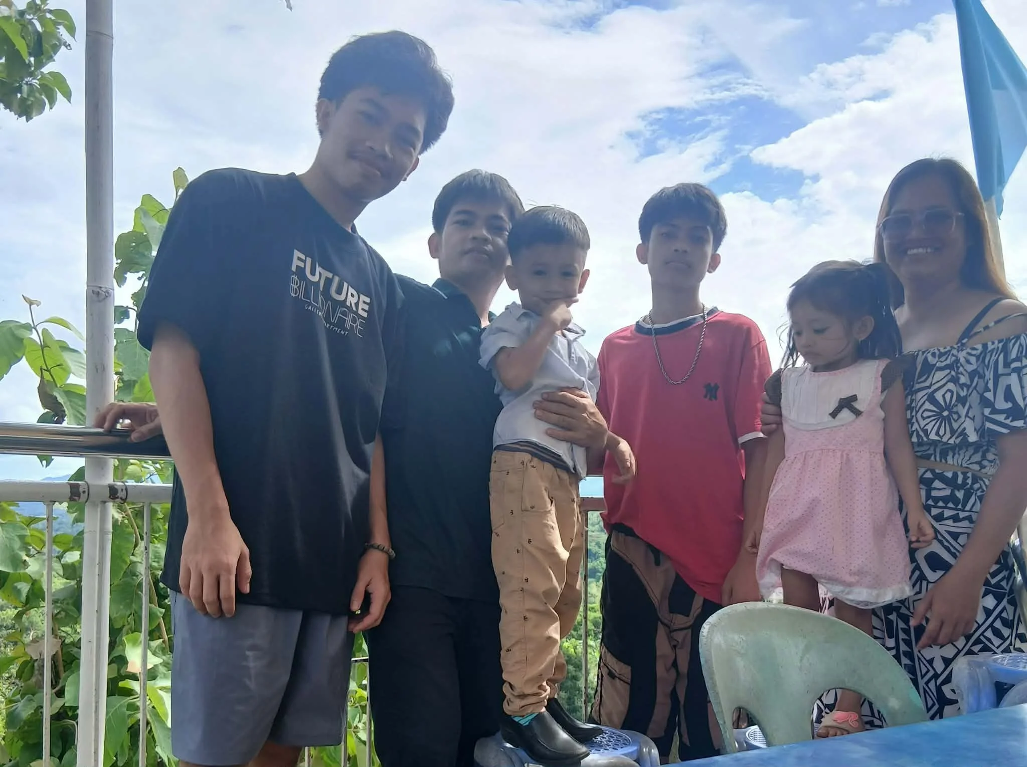 A group of six people, five children and one adult woman, standing outdoors on a cloudy day. They are on a balcony or terrace with a railing, and there are green plants and blue sky with clouds in the background. The children are dressed casually, an