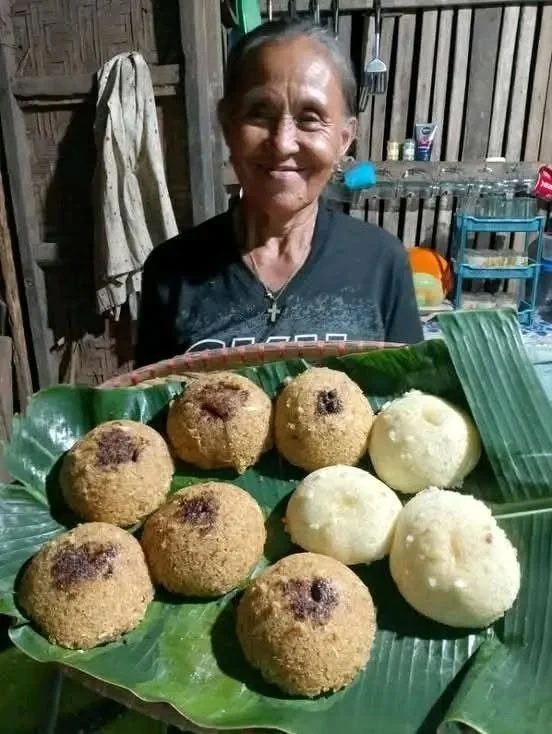 An elderly woman smiling behind a tray of eight round, fried food items on green banana leaves in a rustic kitchen setting.