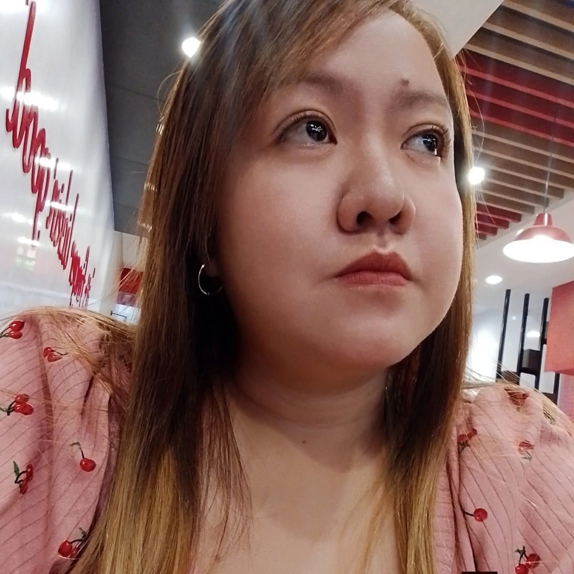 Close-up of a young woman with long reddish-brown hair, wearing hoop earrings, in a setting with a white wall and ceiling with wooden beams. She's wearing a pink top with red cherry patterns, and the photo focuses on her face with a neutral expressio