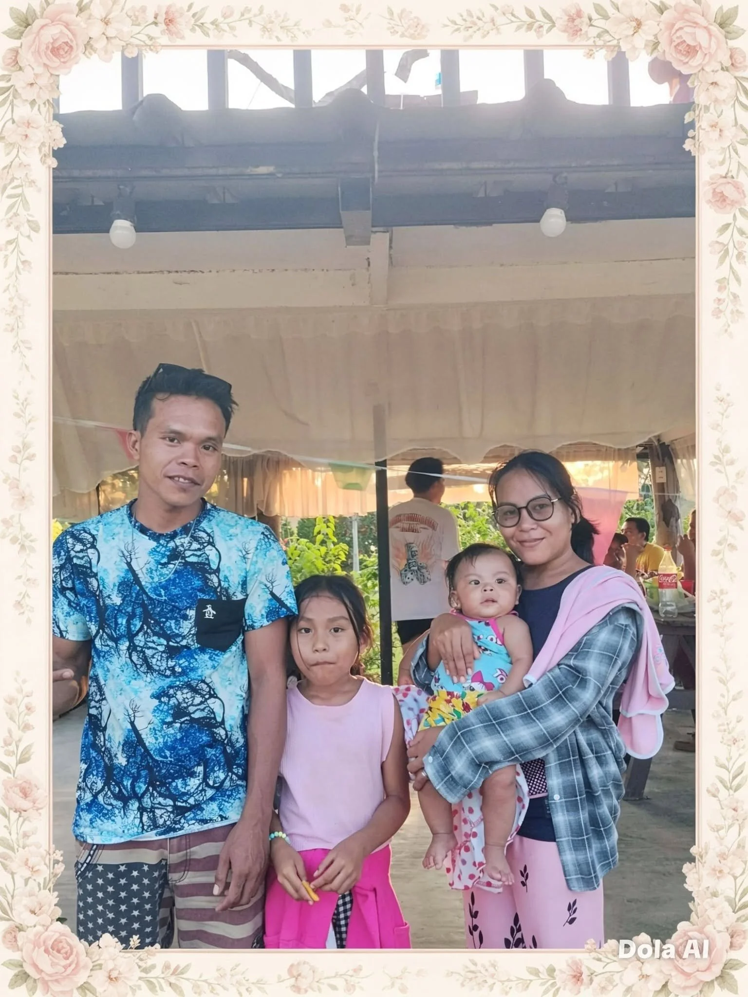 A family portrait featuring a man, woman, young girl, and baby girl posing outdoors under a covered area with other people in the background. The man is wearing a blue patterned shirt, the woman is wearing pink pajamas and glasses, the girl is wearin