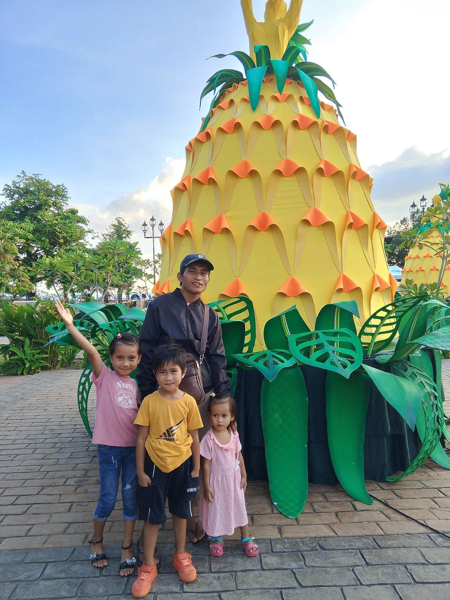 A group of five children and one adult standing in front of a large decorative pineapple prop at an outdoor setting during daytime. The children are smiling and looking at the camera, with the background showing trees, lampposts, and a partly cloudy 