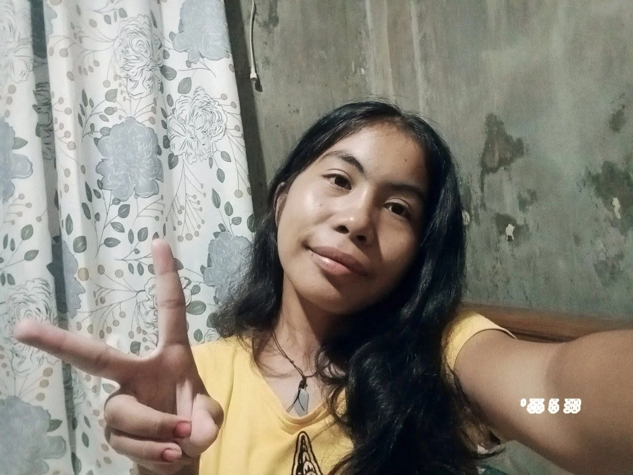 A young woman with long black hair is taking a selfie indoors, making a peace sign with her right hand, wearing a yellow t-shirt and necklace, with a floral curtain and a concrete wall in the background.