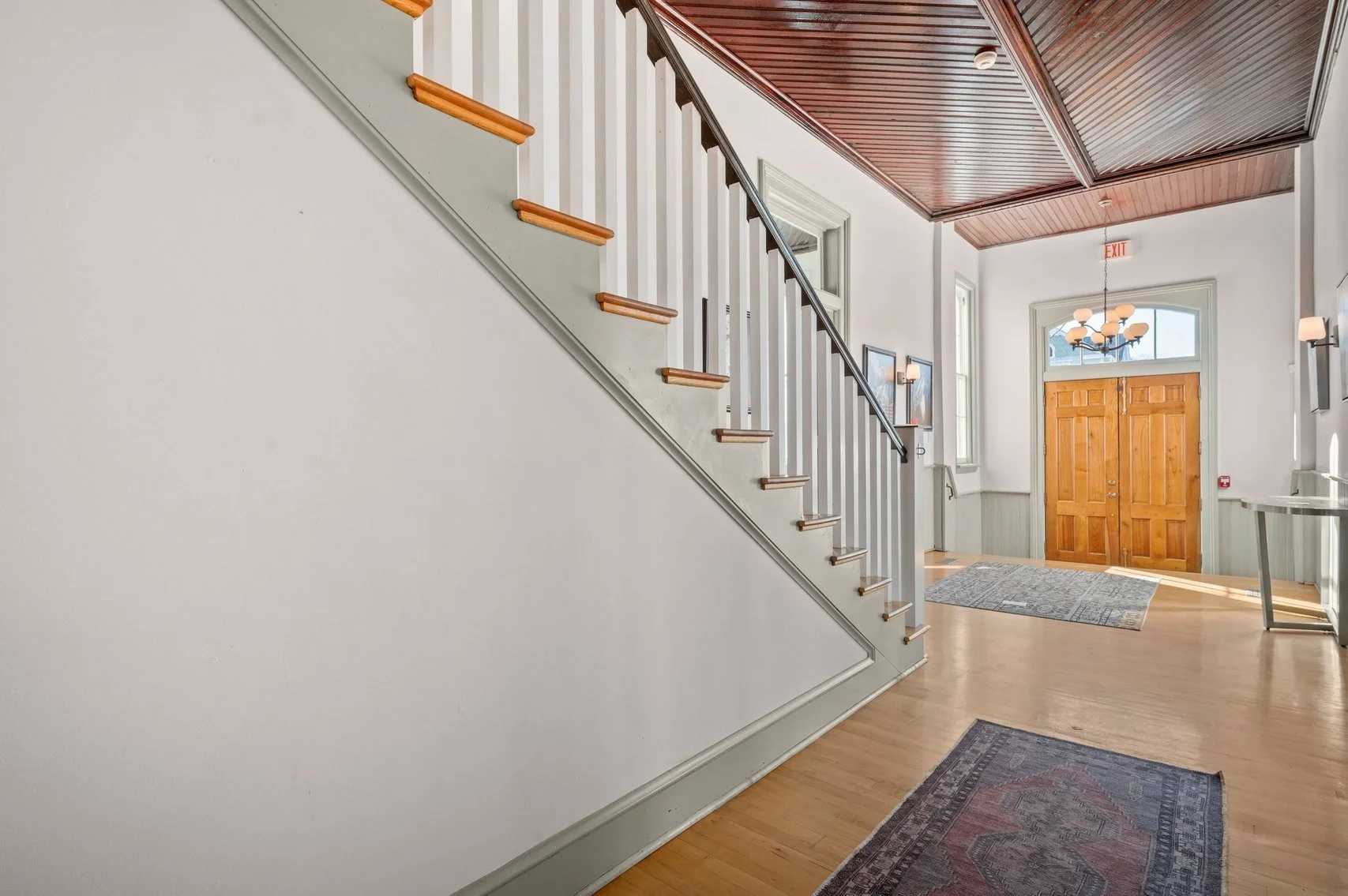 Bright entrance hallway with wooden front door, geometric ceiling, hardwood floors, area rugs, and staircase with white railing and wooden steps.