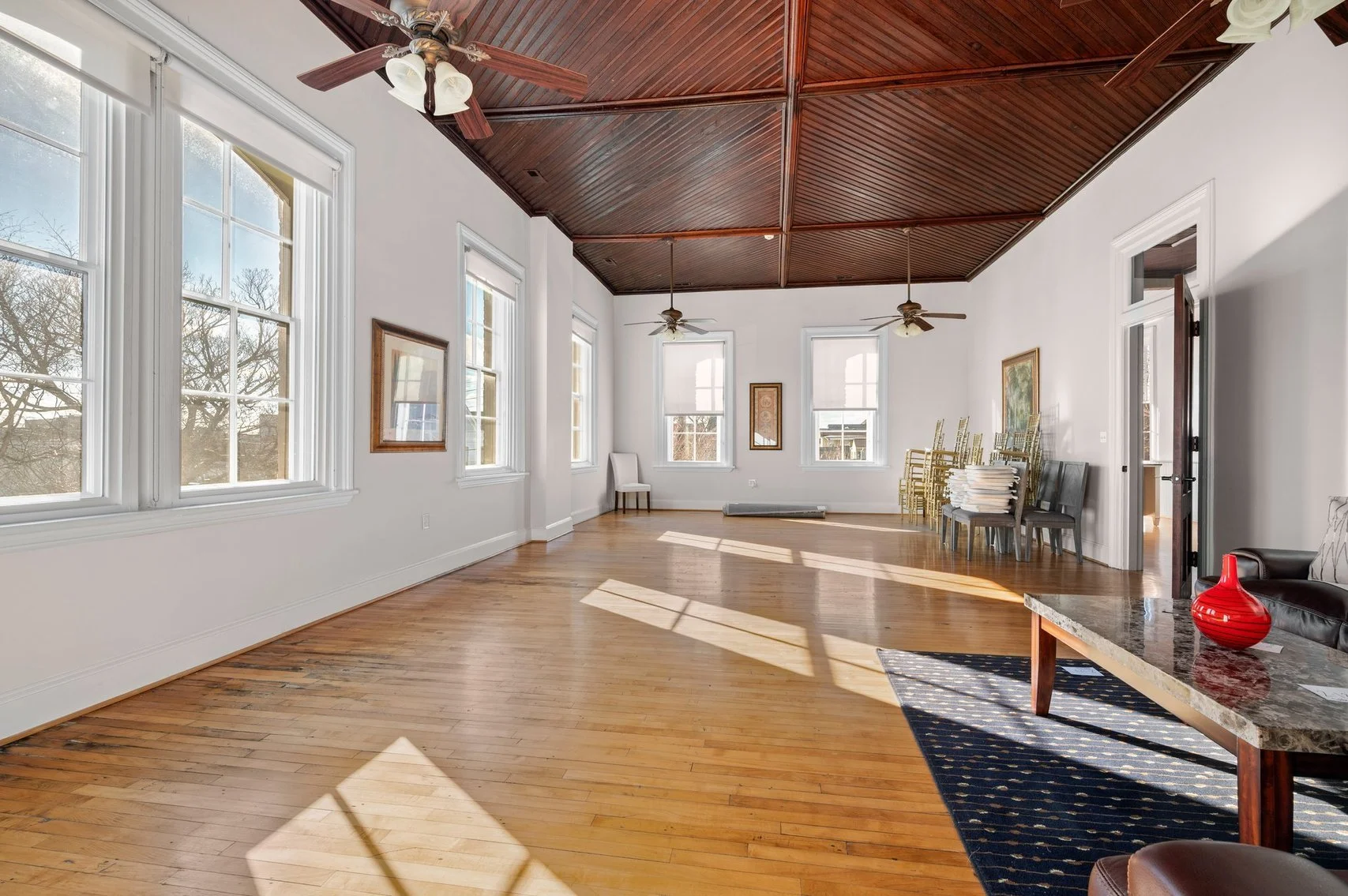 Empty room with hardwood floors, white walls, large windows, and a dark wood vaulted ceiling with ceiling fans.