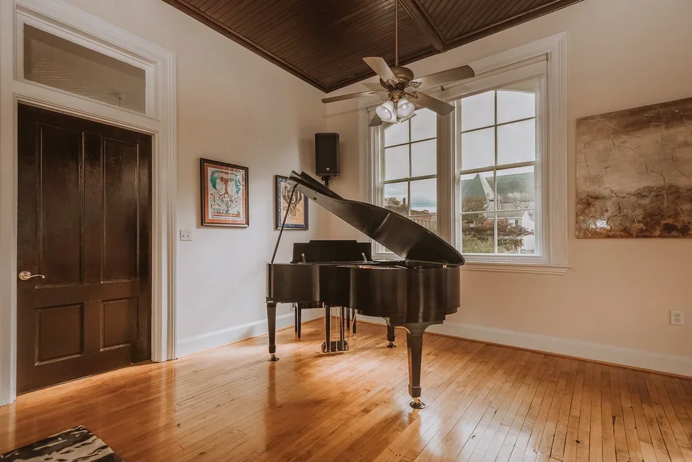 A black grand piano in a room with large windows, hardwood floor, ceiling fan, and artwork on the wall.