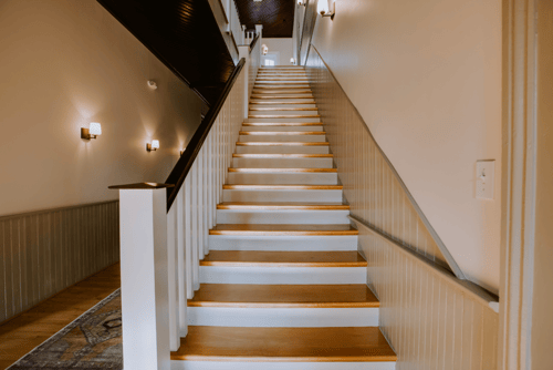 Indoor staircase with wooden steps, white risers, and a black handrail on the right side, leading to an upper floor, with wall-mounted lights along the left wall.