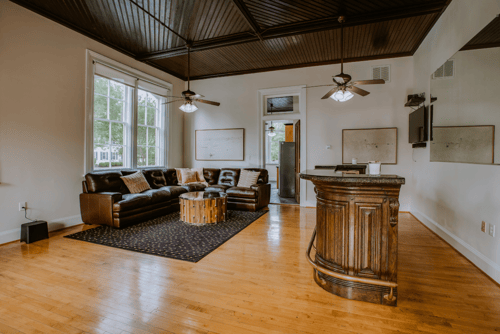 Living room with a brown leather sofa, a small round wooden coffee table, large windows, and a wooden bar counter.