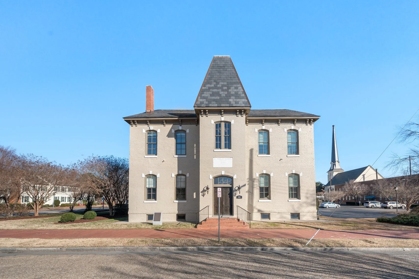 A historic three-story brick historic school building with a steep pointed roof and arched windows, featuring a central entrance with stairs, a small lawn area, and a sidewalk in front, against a clear blue sky with a church steeple and nearby trees.