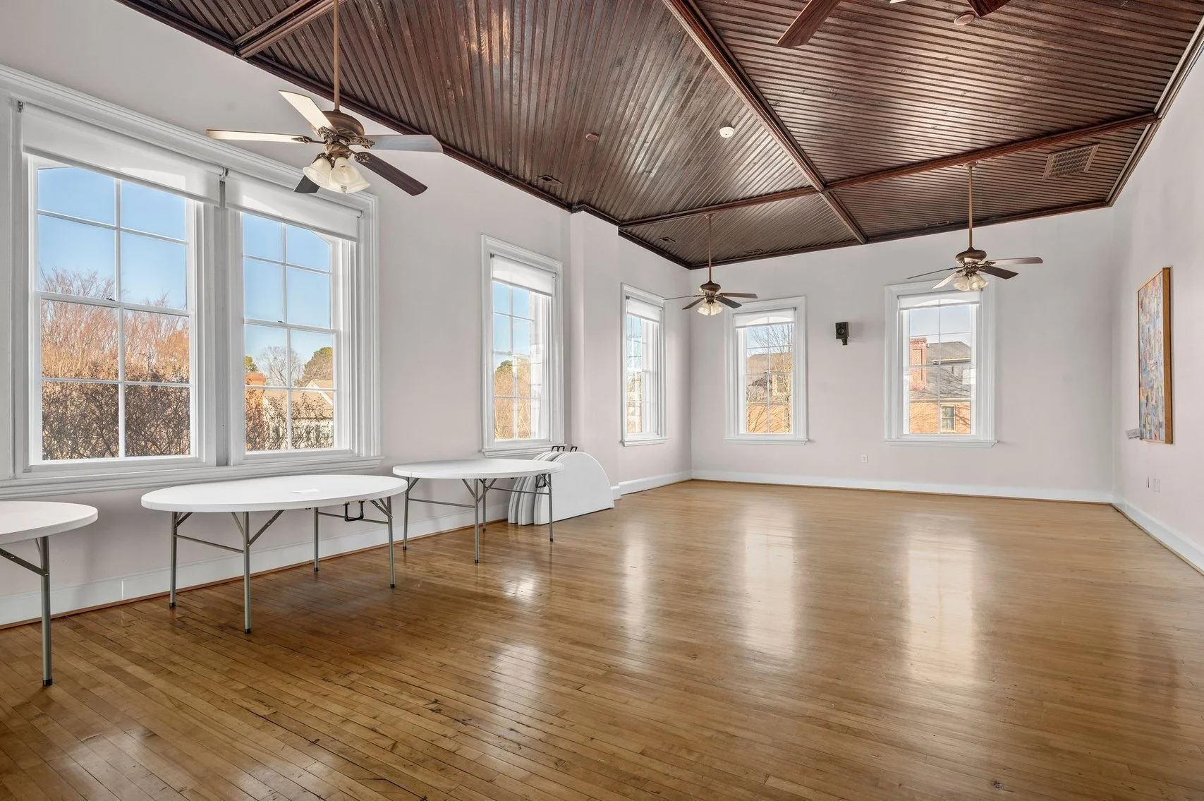 Empty room with hardwood floors, white walls, large windows, ceiling fans, and a decorative wood-paneled ceiling