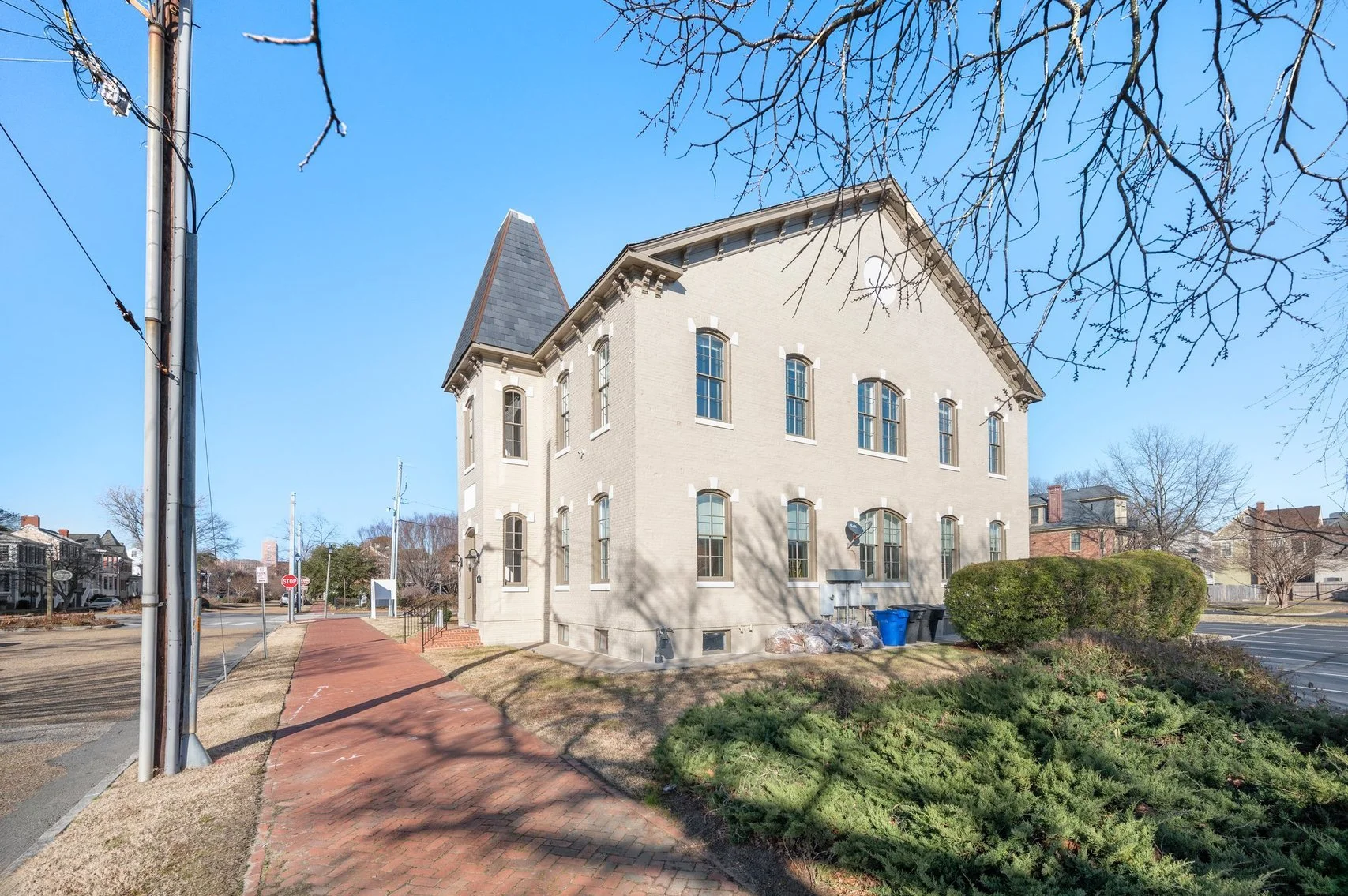 A large, historic light-colored brick building with a turret and arched windows, situated on a corner with a brick sidewalk, trees, and a parking lot under a clear blue sky.