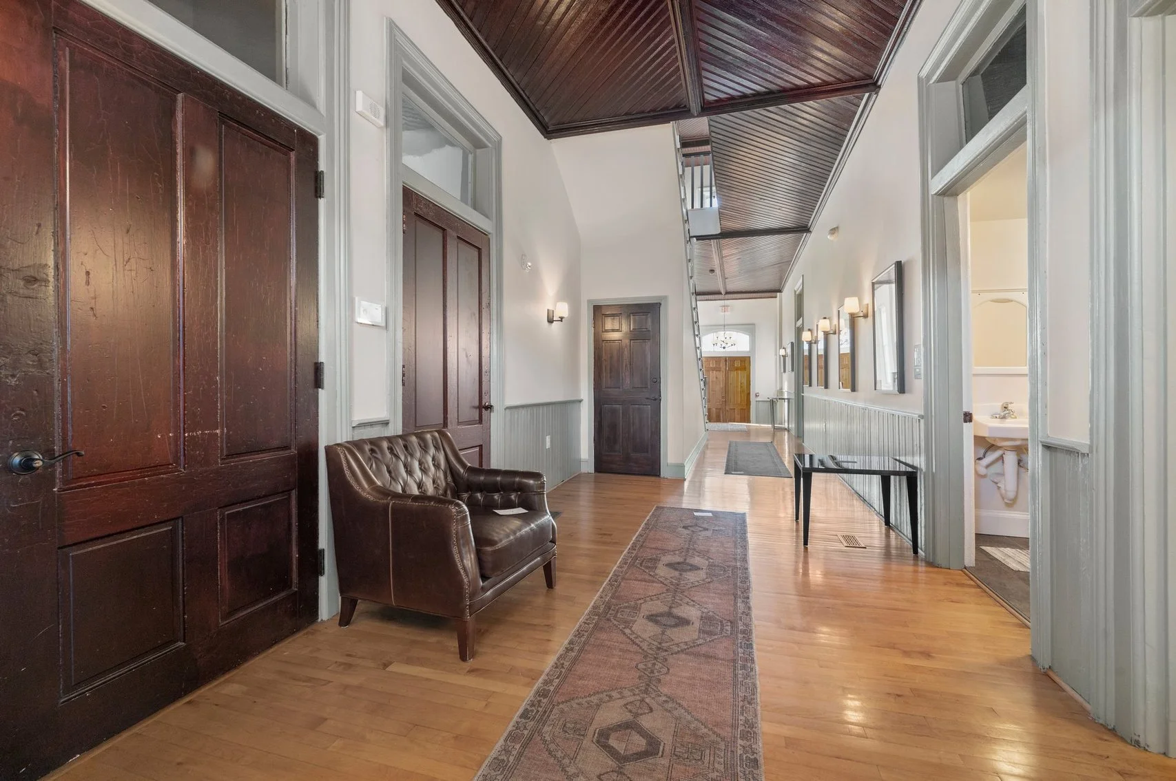 An entry hallway with hardwood floors, vintage rugs, white walls, and wooden ceiling panels, featuring a brown leather bench on the left, several wall lamps, and multiple doors leading to different rooms.