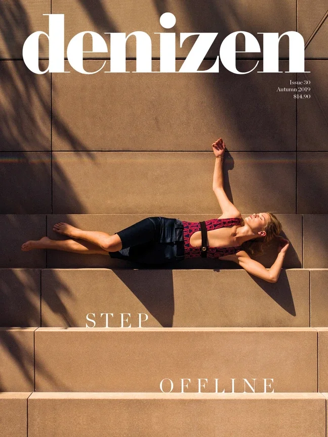 A woman lies on sideways on outdoor stairs in sunlight, wearing a stylish black and red outfit, with her eyes closed and one arm raised above her head.