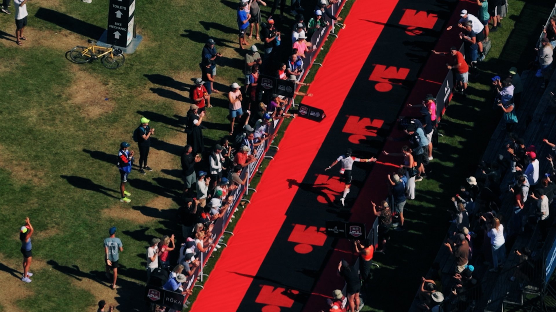 Crowd of people lining a red and black race finish line with logos, some taking photos, in an outdoor sports event.