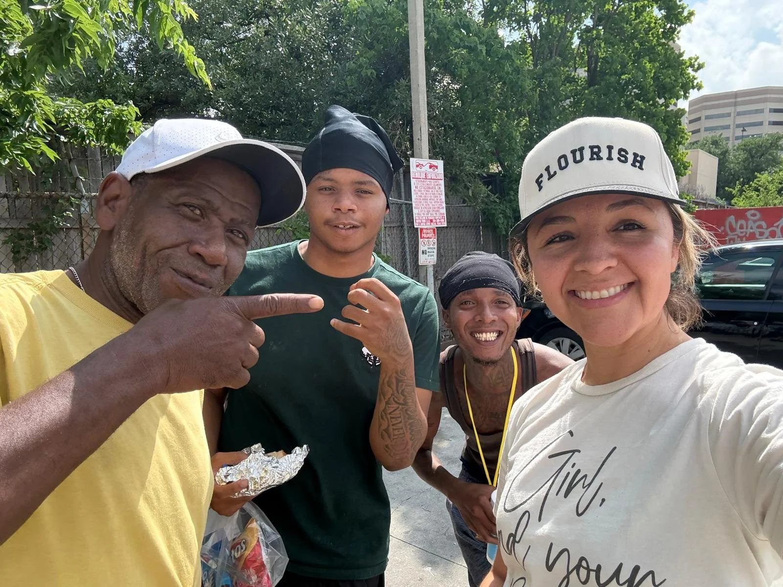 Four people smiling and looking at the camera outdoors, with trees and cars in the background. The group consists of an older man wearing a white cap and yellow shirt, a woman wearing a white cap and a white shirt, and two men in black bandanas and dark shirts. One of the men is pointing downward while holding an foil-wrapped item.