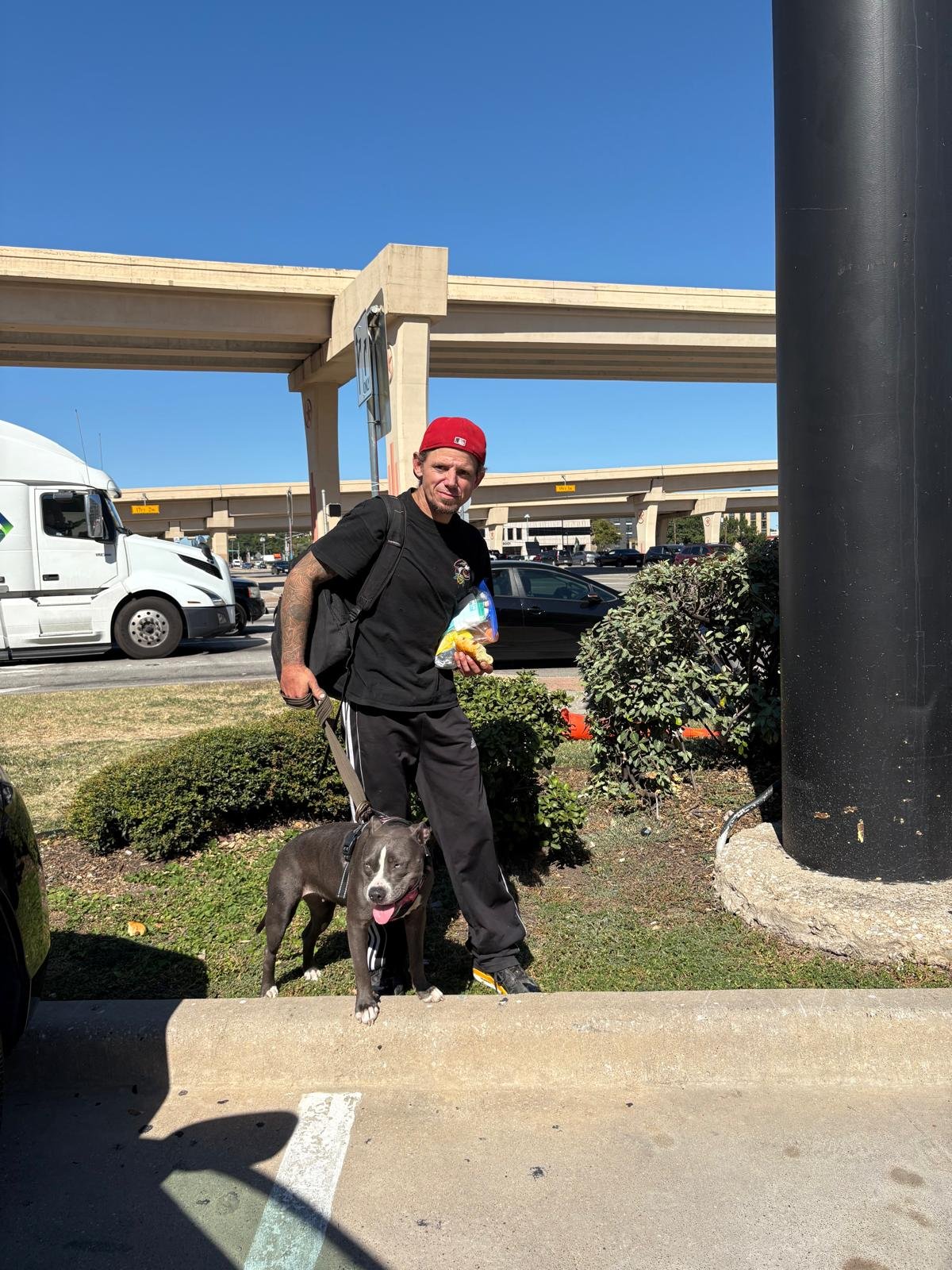 A man with a red cap, black t-shirt, in black pants and sneakers stands beside a black and white dog in a parking lot under a highway overpass. He is holding a bag of snacks and a drink, and there are cars and trucks in the background.