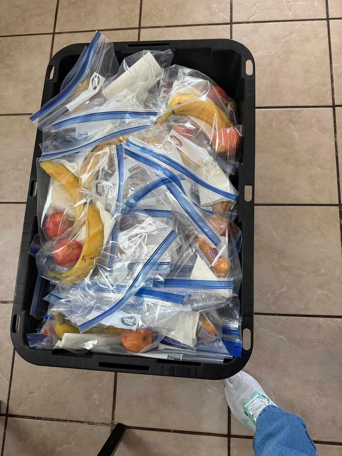 A black plastic tray filled with sealed plastic bags of sliced bananas, apples, and other fruits, placed on a tiled floor. The photo is taken from above, showing a person wearing blue jeans and white sneakers standing nearby.