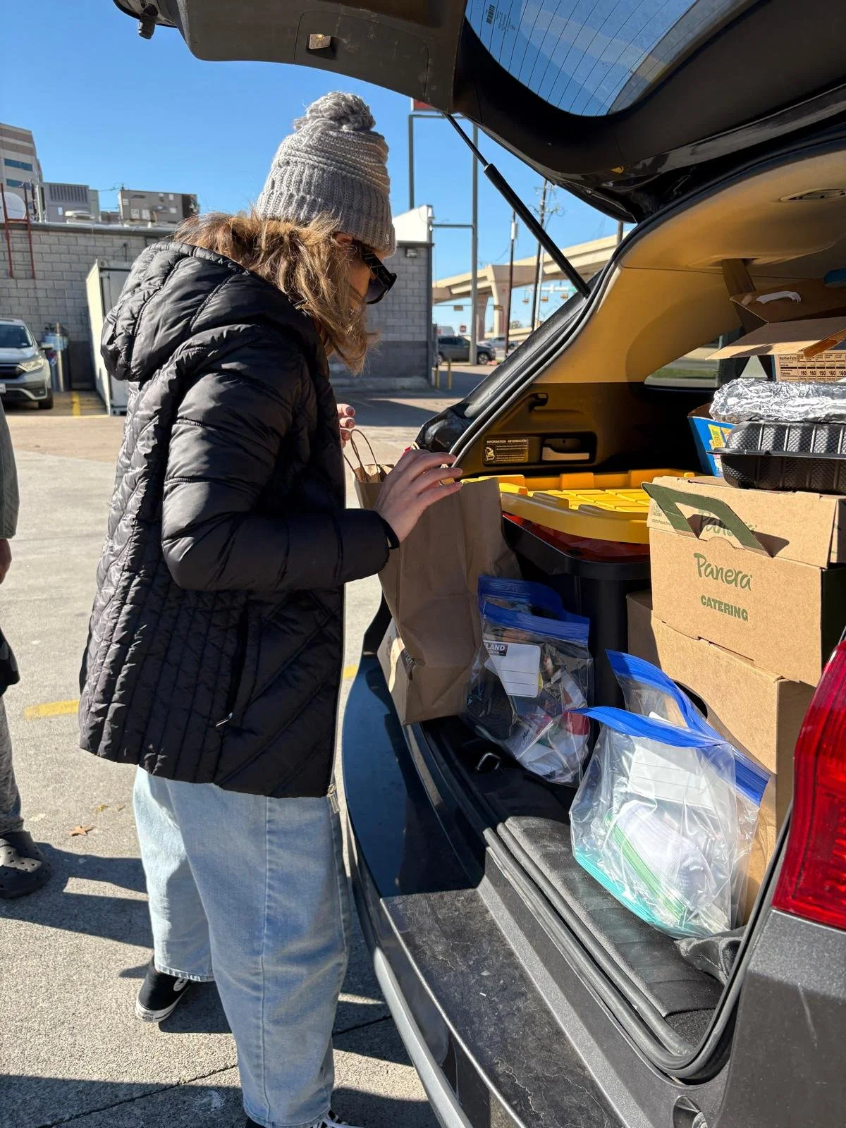 A woman in a black puffer jacket, gray beanie, and sunglasses loading groceries into the trunk of a black SUV in a parking lot.